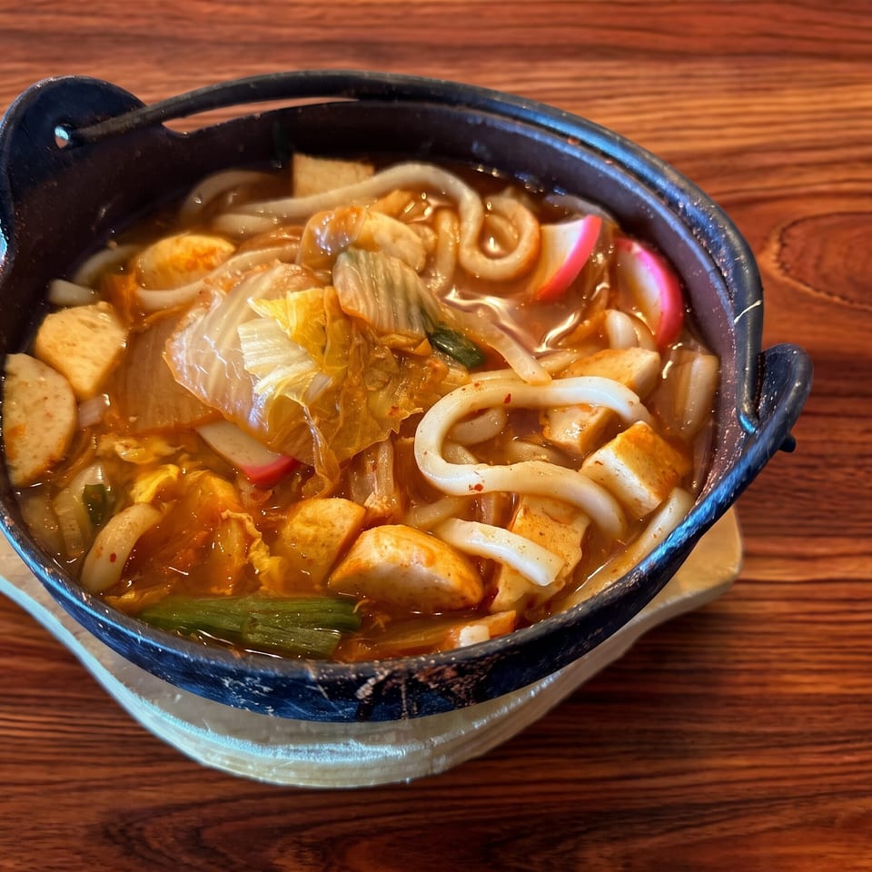 A black pot of spicy udon noodle soup with kimchi, tofu, and fish cakes on a wooden table.